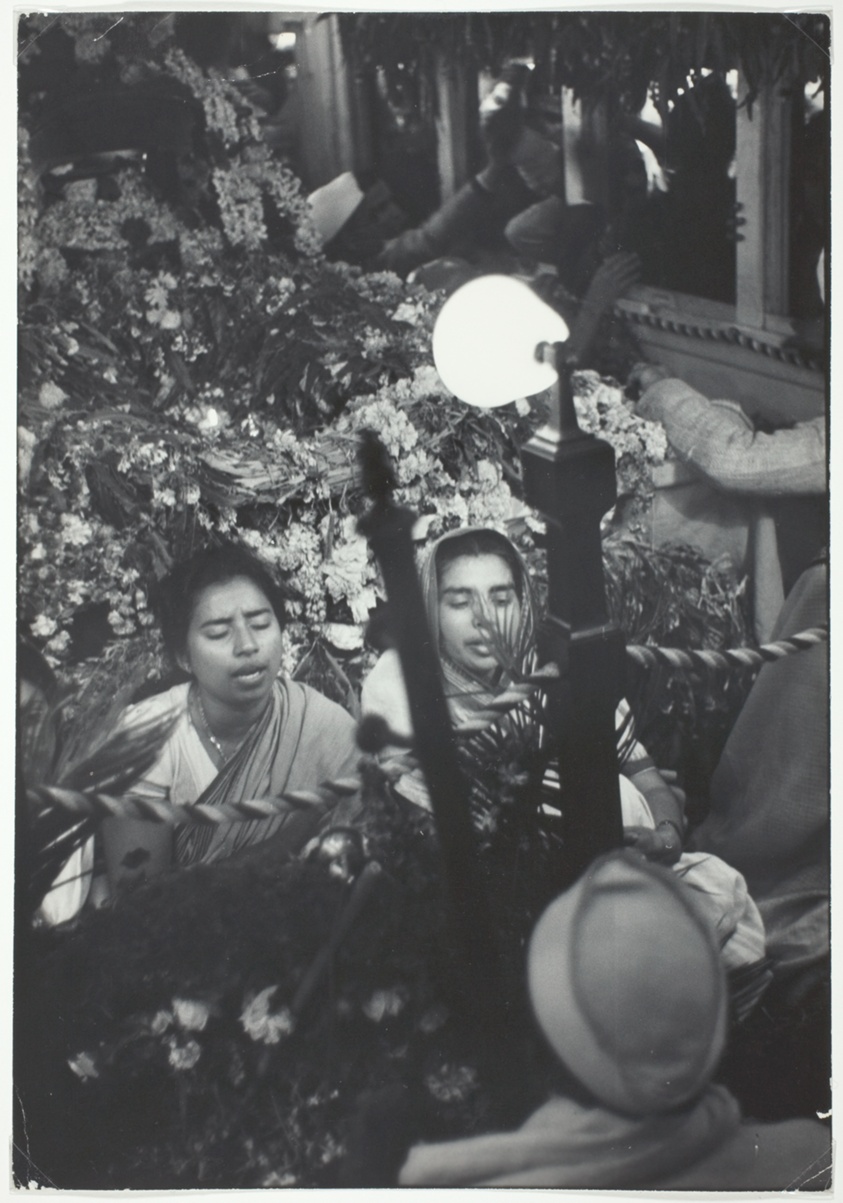Two Women Sit Reciting Prayers, Facing Light with Mass of Flowers